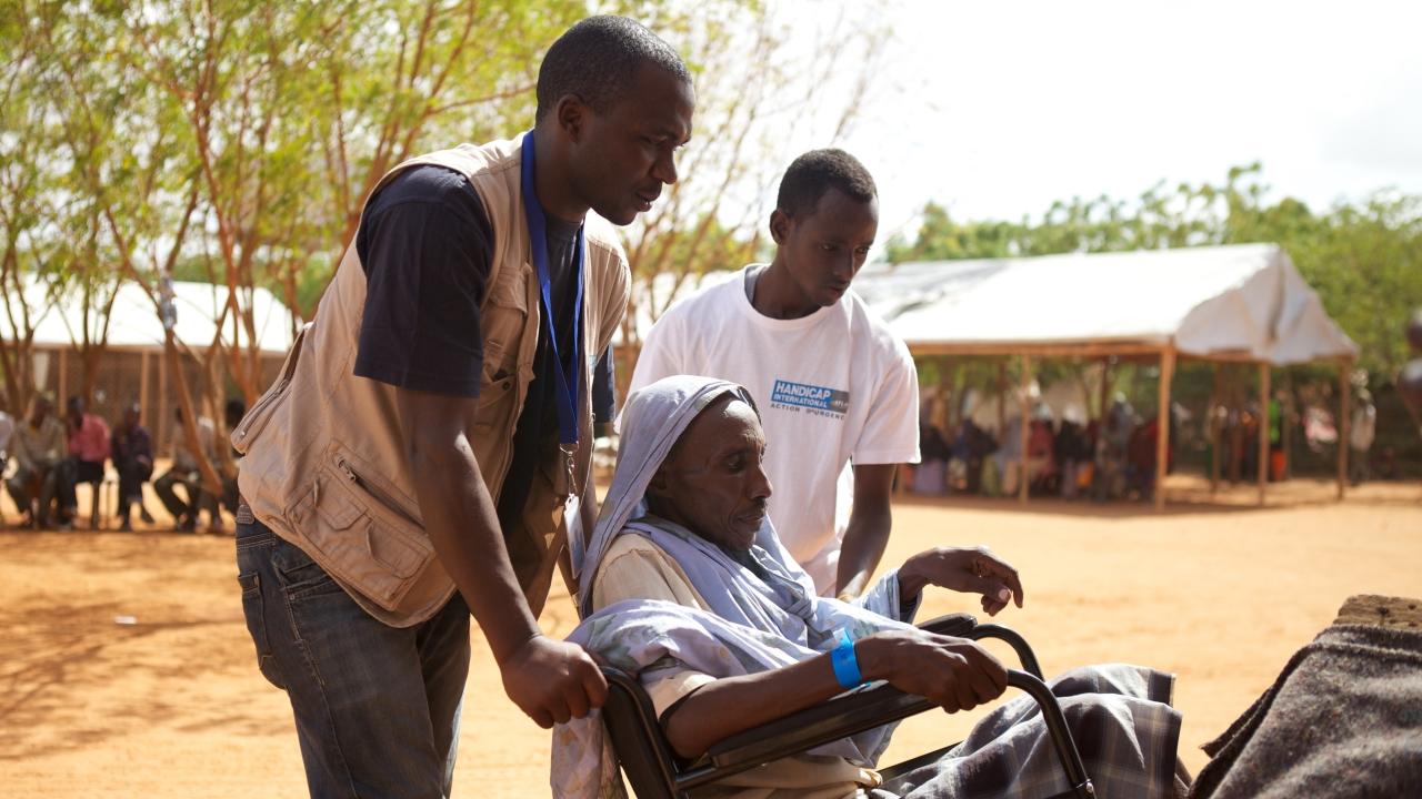 Zwei Mitarbeiter von Handicap International schieben einen Mann im Rollstuhl