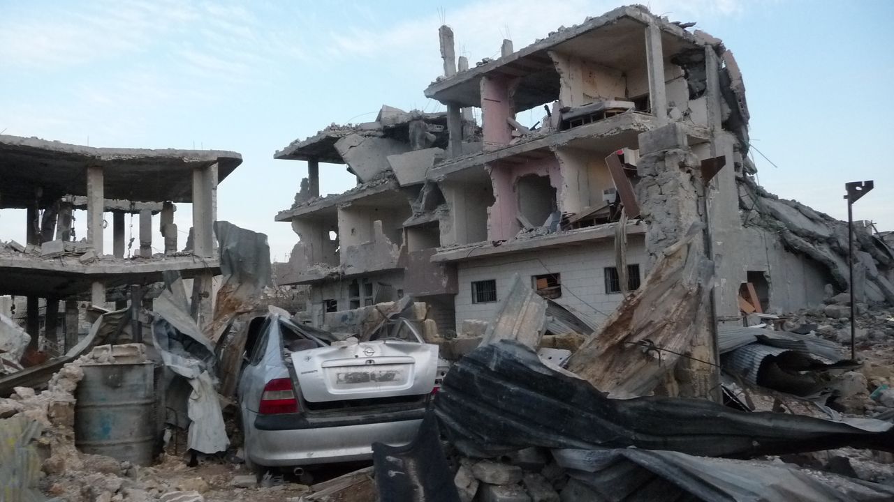 Ein Bombenkrater in der Stadt Kobane in Syrien. Im Hintergrund sind zwei umgedrehte Autos.
