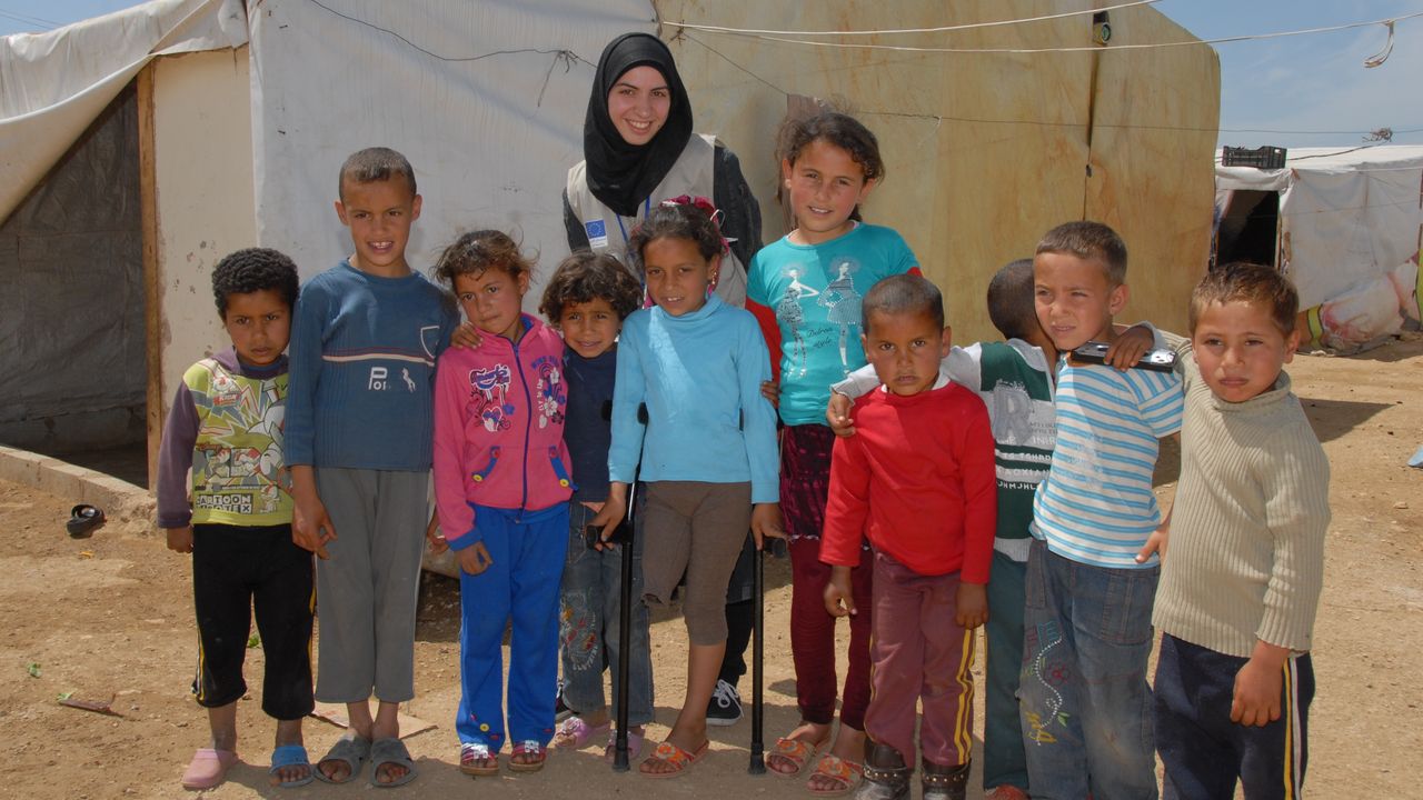 Ahlam steht auf Krücken im Camp, umring von Kindern und einer Helferin von Handicap International