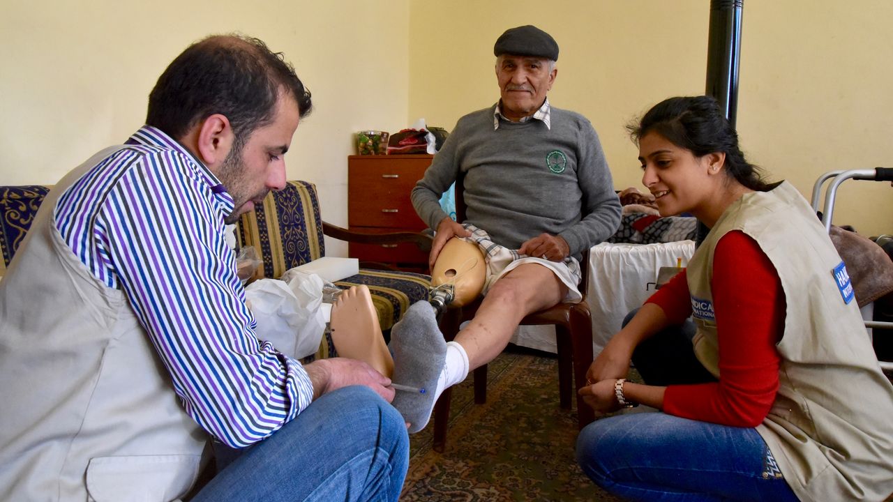 Mamdouh sitzt auf einem Stuhl. Vor ihm knien der Orthopädiemechaniker und die Physiotherapeutin und untersuchen sein Bein samt Prothese.
