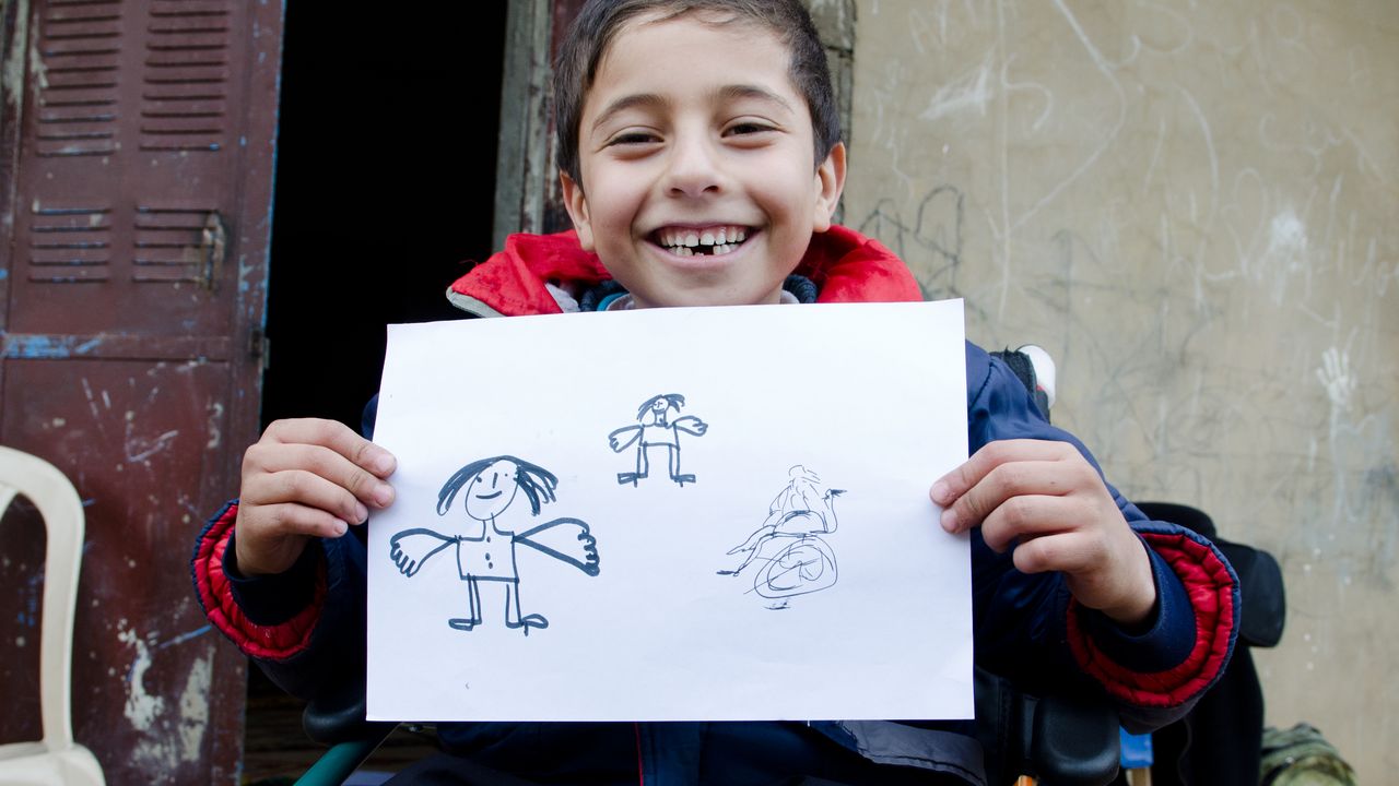 der siebenjährige Ahmad aus Syrien sitzt im Rollstuhl und zeigt der Kamera stolz seine Zeichnungen von Engeln