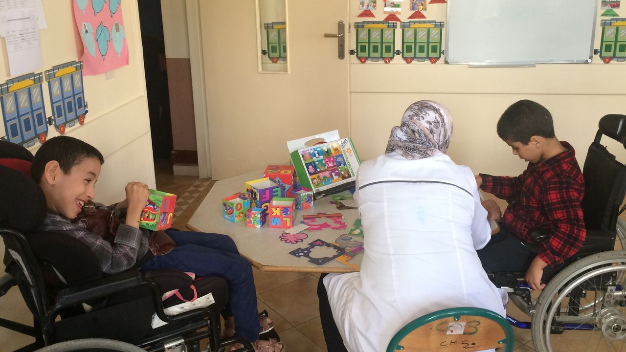 Projektbesuch in Marokko. Zwei Jungen im Rollstuhl sitzen sich an einem Tisch gegenüber. Der Linke spielt mit einem großen bunten Würfel. Mit dem rechten Jungen beschäftigt sich eine Frau, die zwischen den beiden Jungen sitzt.