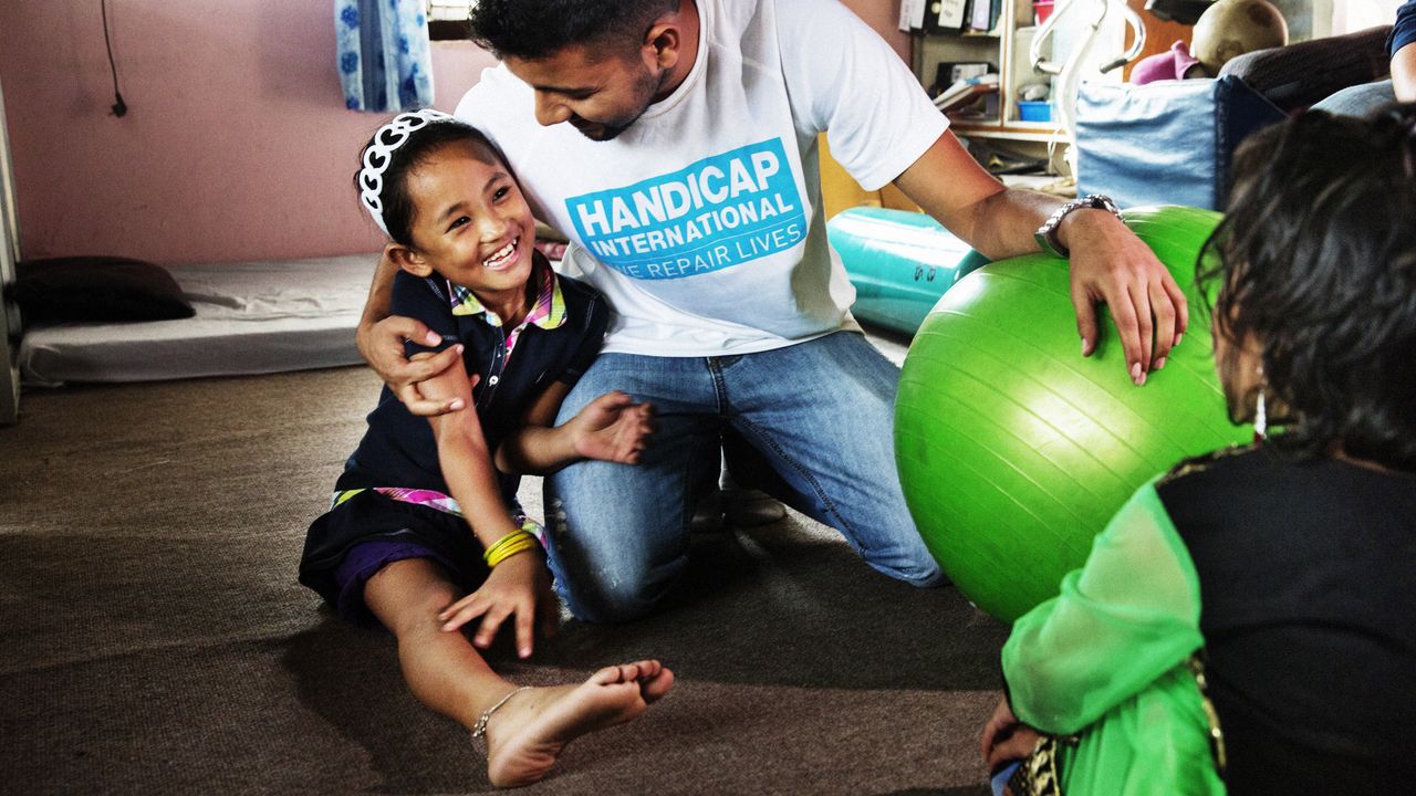 Ein Physiotherapeut von Handicap International sitzt mit dem nepalesischen Mädchen Nirmala auf dem Boden und hält sie im Arm, während sie sich vertraut anlachen.