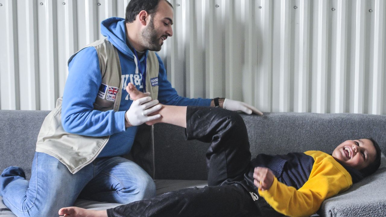Abdel Rahman liegt auf auf einer Couch. Sein Physiotherapeut von Handicap International kniet vor ihm und hält mit einer Hand Abdel Rahmans linkes Bein, dass dieser angewinkelt hat.