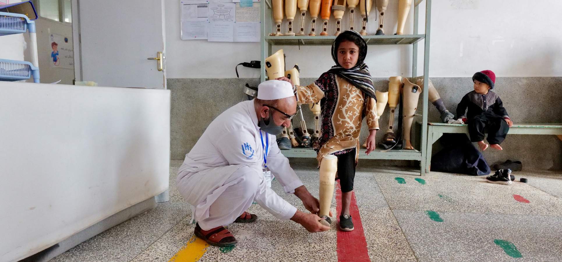 Ein junges Mädchen bekommt von einem Experten von HI eine Prothese angepasst. Sie stehen in einem klinischen Untersuchungsraum, im Hintergrund ist ein Regal mit Prothesen zu sehen.