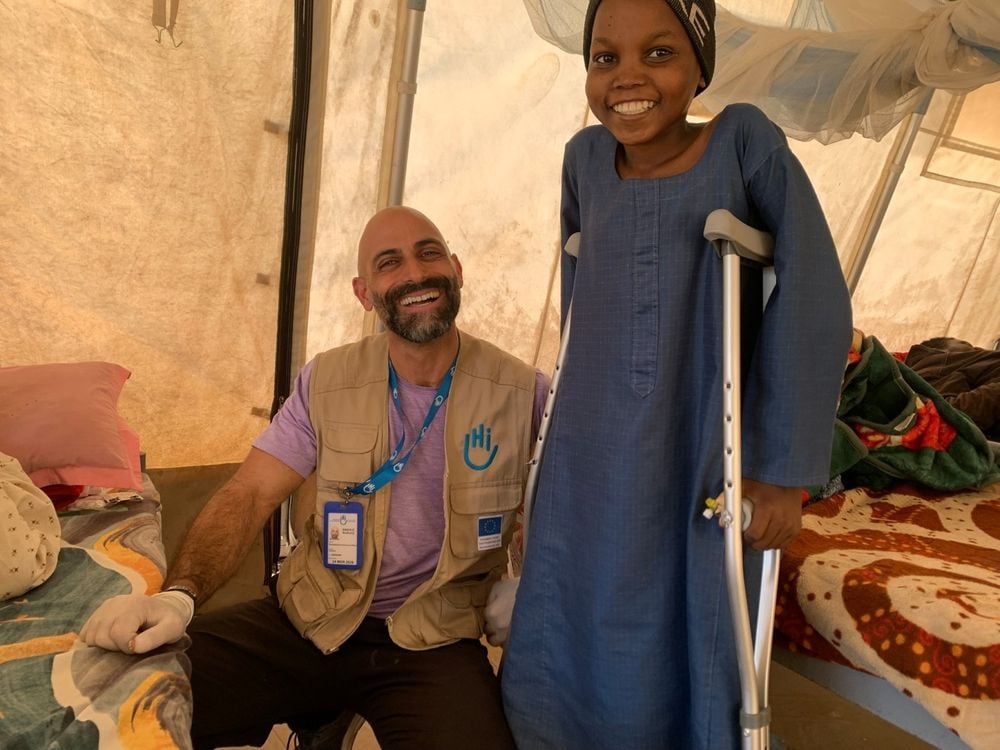 Ein Junge mit Krücken mit seinem Physiotherapeuten im Hintergrund. Beide lächeln in die Kamera