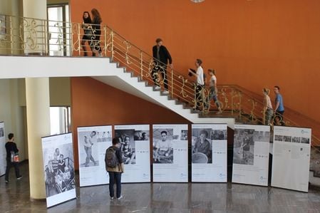 Ein Foyer einer Universität. Menschen gehen eine Treppe hinauf. Vor ihnen stehen Roll-ups einer Ausstellung. Ein Mann bleibt davor stehen und betrachtet die Ausstellung. 