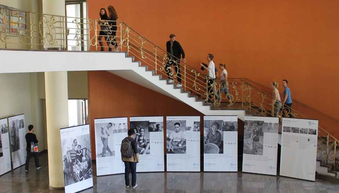 Im Foyer steht eine Ausstellung. Dahinter befindet sich eine Treppe, auf der sich gerade Menschen befinden. 
