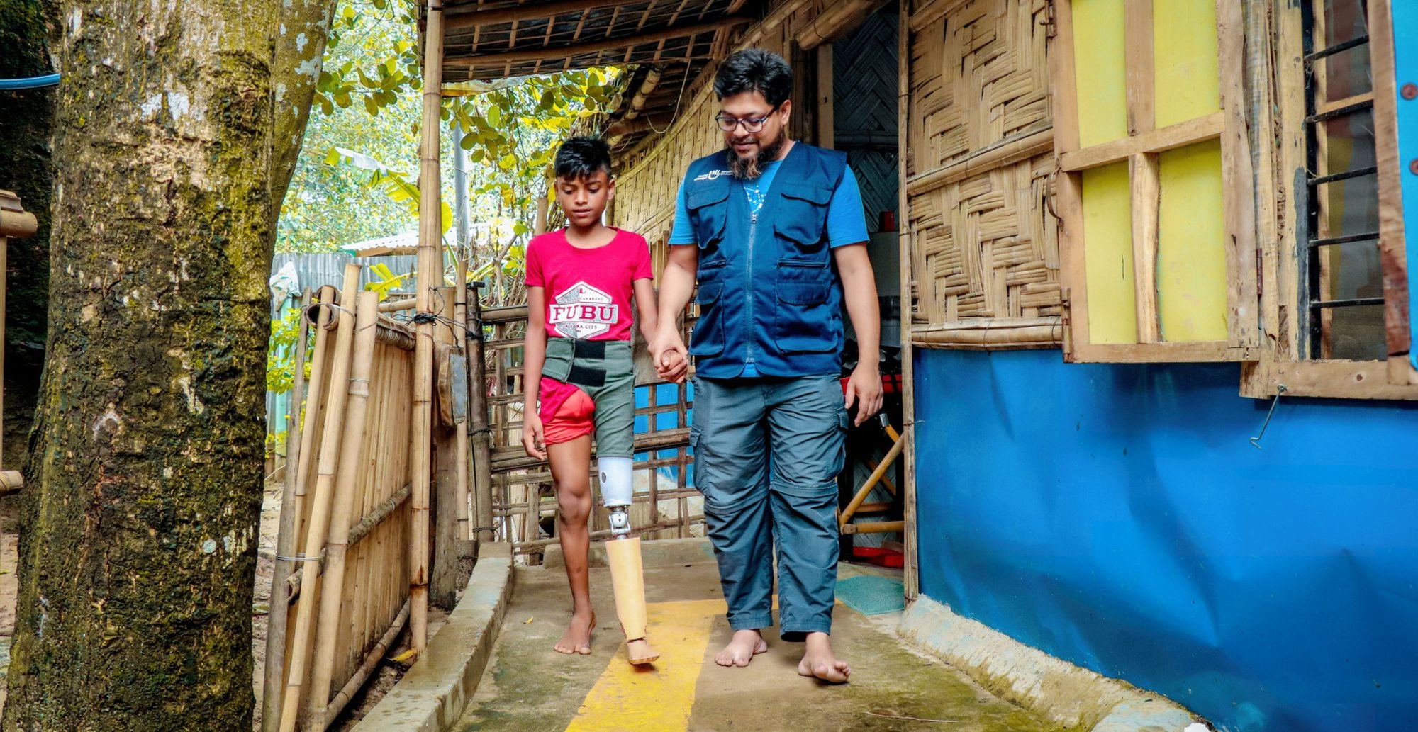 Junge aus Bangladesch mit Beinprothese wird von seinem Physiotherapeuten an der Hand geführt.