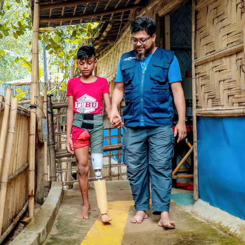 Junge aus Bangladesch mit Beinprothese wird von seinem Physiotherapeuten an der Hand geführt.