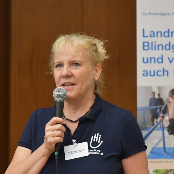 Eine Frau mit blonden Haaren trägt ein dunkelblaues Poloshirt mit Handicap International Logo und spricht in ein Mikrofon.