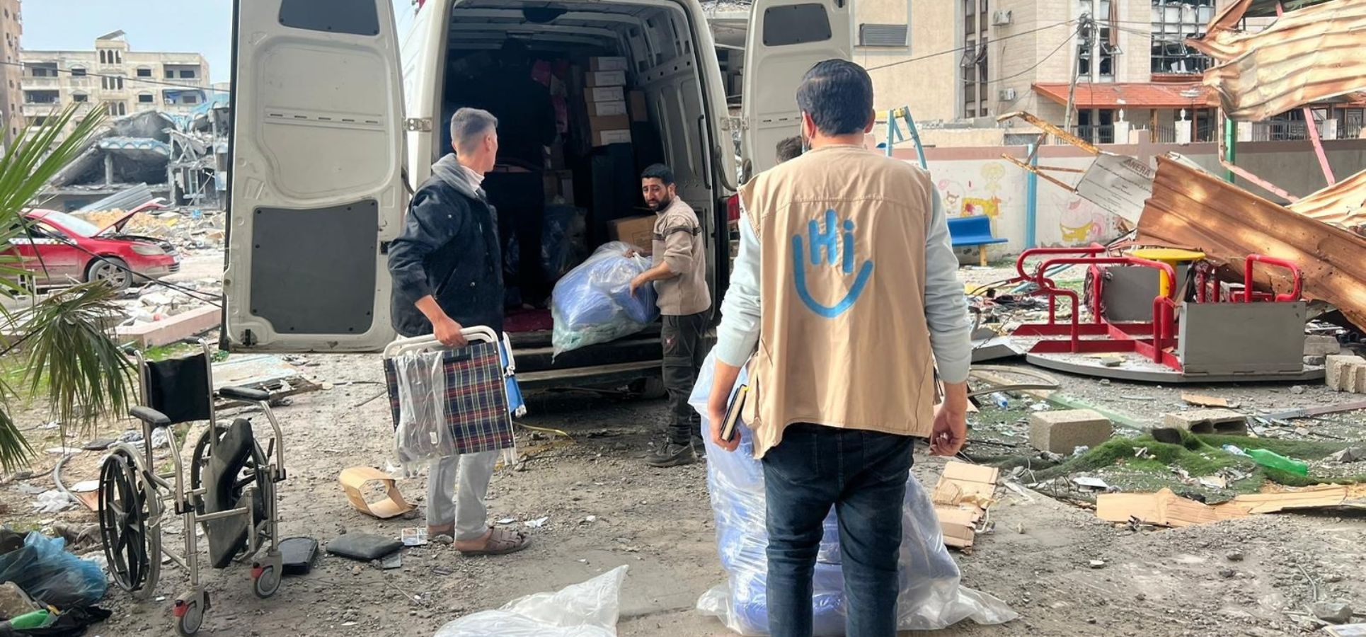 Unsere Team laden Rollstühle, Krücken, etc. aus einem Transporter vor einem Krankenhaus. Im Hintegrund liegt die Stadt in Trümmern.