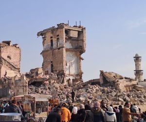 Häuser, die durch Bomben zerstört wurden. Vorne stehen viele Menschen auf einem Platz.