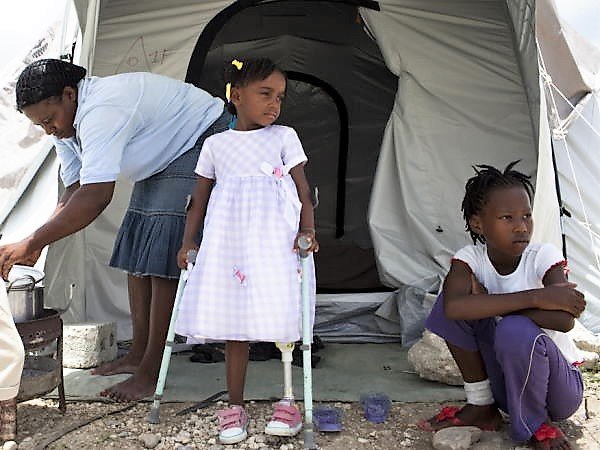 Ein kleines Mädchen, ein Junge und eine Frau aus Haiti vor einer Notbehausung. Das Mädchen hat ein prostethisches Bein. © William Daniels / HI