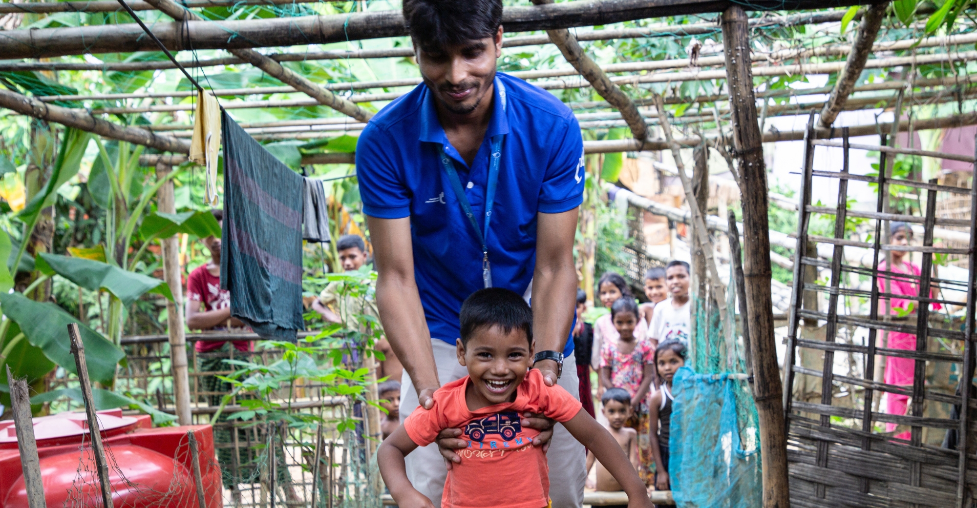 Ein kleiner Junge läuft und lächelt strahlend. Unter den Armen hält ihn ein HI-Helfer in blauen Klamotten. Im Hintergrund schauen viele Kinder dabei zu. 