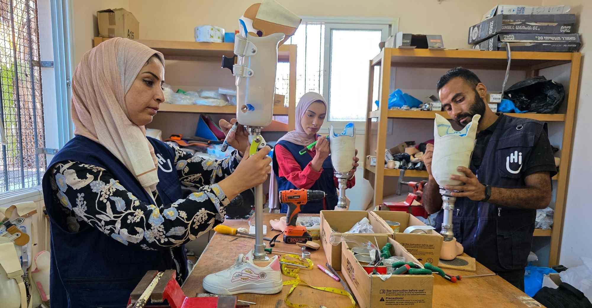 Zwei Mitarbeiterinnen und ein Mitarbeiter von Handicap International stehen an einem Tisch und bauen Beinprothesen. 