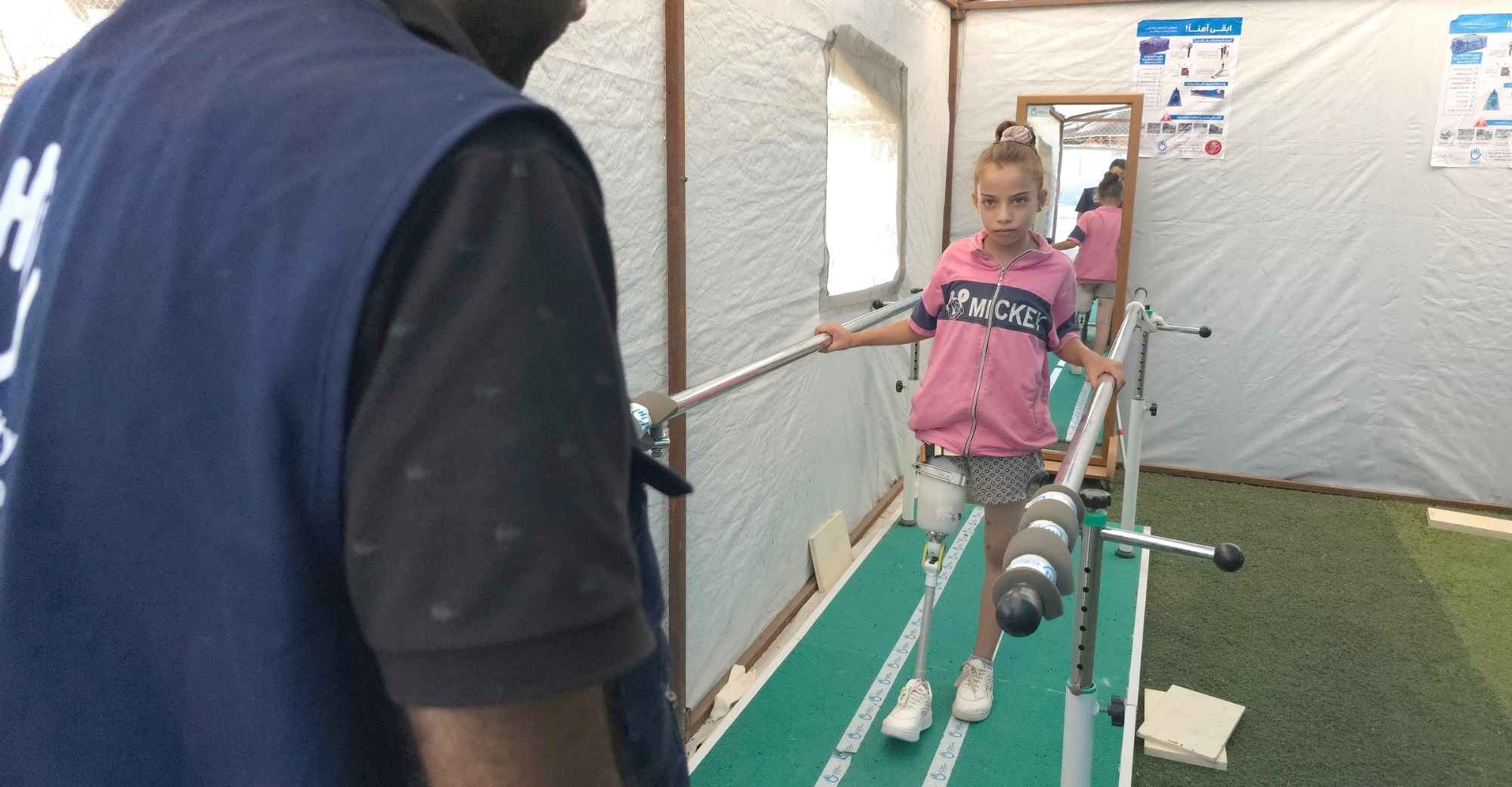 Ein Mädchen mit Beinamputation übt das Laufen mit einer Prothese an einer Parallelstange. Vor ihr steht ein Helfer von Handicap International. 