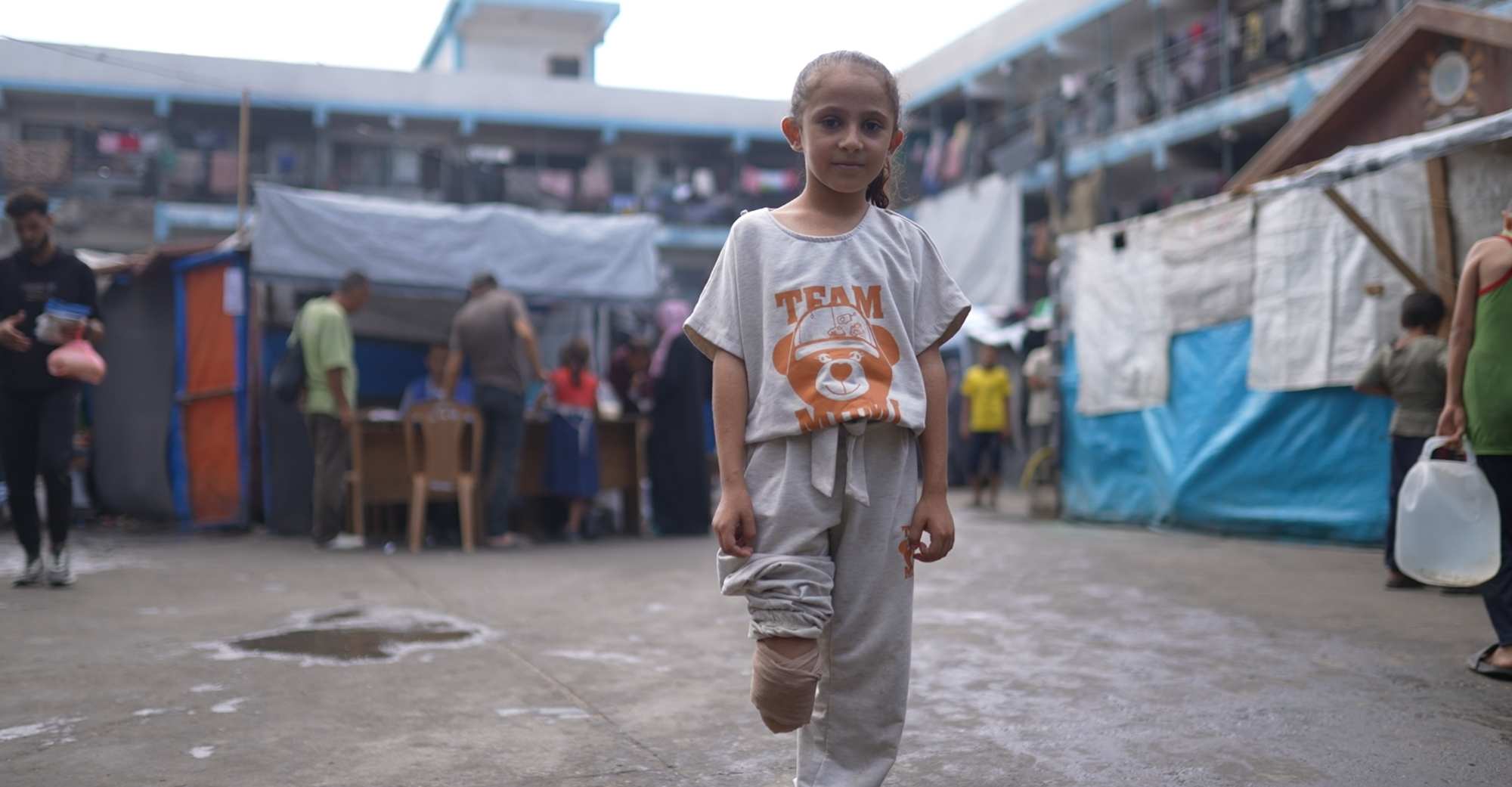 Ein Mädchen mit Beinamputation steht auf einem großem Hof. Um sie herum sind einige Menschen. 