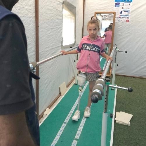 Ein Mädchen mit Beinamputation übt das Laufen mit einer Prothese an einer Parallelstange. Vor ihr steht ein Helfer von Handicap International. 