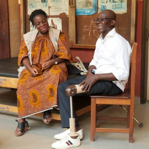 Ein Mann mit einer Beinprothese sitzt auf einem Stuhl und lächelt. Neben ihm sitzt eine Mitarbeiterin von HI in einem bunten Kleid und Weste und lächelt auch und schaut auf seine Prothese.