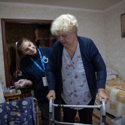 Eine ältere Frau mit kurzen blonden Haaren stützt sich auf ihren Rollator. Hinter ihr steht eine Helferin von Handicap International und lächelt sie an. Im Hintergrund ist ein Wohnzimmer.