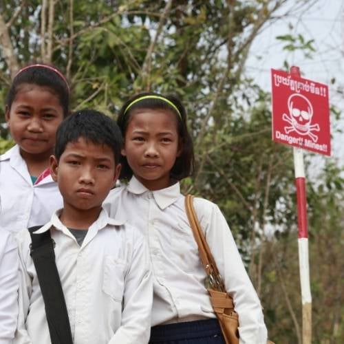 Eine kleine Gruppe Schulkinder steht links und schaut ernst. Rechts neben ihnen steht ein Schild, das mit Totenkopf-Symbol vor Minen warnt.