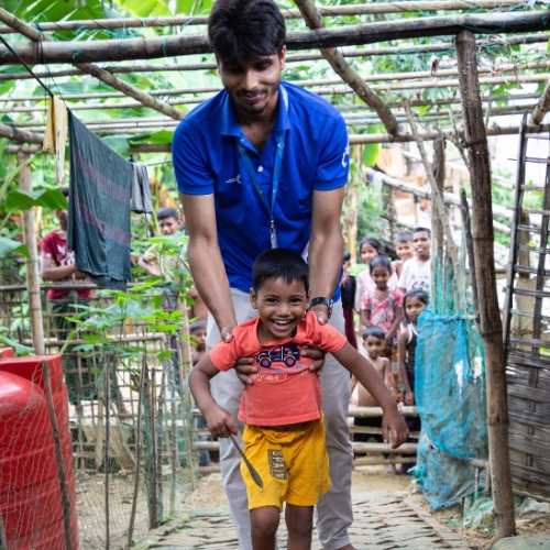 Ein kleiner Junge läuft und lächelt strahlend. Unter den Armen hält ihn ein HI-Helfer in blauen Klamotten. Im Hintergrund schauen viele Kinder dabei zu. 
