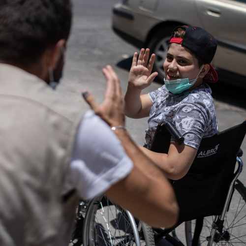 Ein Junge aus Syrien sitzt lächelt in einem Rollstuhl und winkt in die Kamera.
