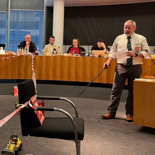 Ein Mann zeigt mit einem Stock auf Minenatrappen. Herum sind Absperrbänder gespannt. Hinter ihm sitzen einige Menschen an einem langen Holzpult und schauen ihm zu.