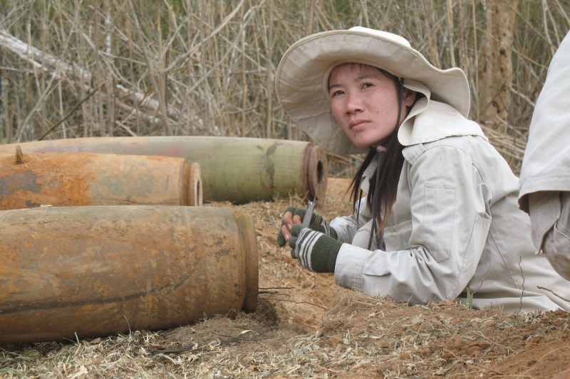 Die weibliche Sprengmeisterin aus Laos schaut ernst in die Kamera. im Hintergrund befinden sich Reste explosiver Waffen.
