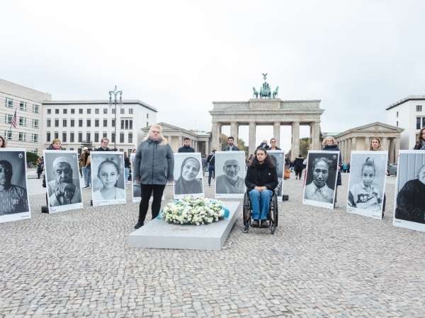 Eine Gruppe von Menschen steht vor dem Brandenburger Tor und hält schwarz-weiße Portraits in den Händen. 