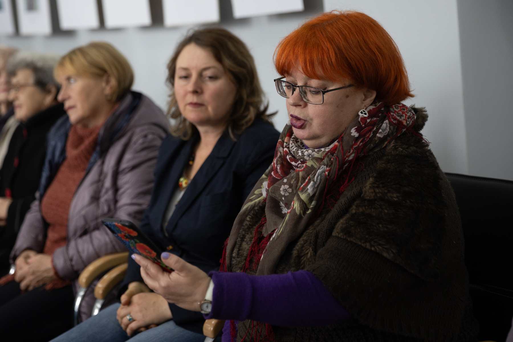 Eine Frau mit kurzen roten Haaren sitzt auf einem Stuhl und spricht. Links sieht man noch mehr Frauen neben ihr sitzen.