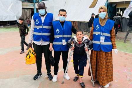 Ein kleiner Junge auf Krücken steht zwischen drei Mitarbeitenden von HI in einem Flüchtlingslager in Gaza.