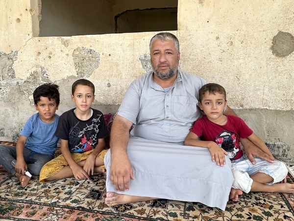 Ein Vater mit seinen drei Söhnen sitzt vor einem Haus mit vielen Einschlagslöchern. 