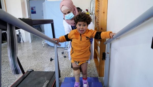 Ein kleines Mädchen mit Prothese geht an Parallelstangen und wird von einer Physiotherapeutin unterstützt.