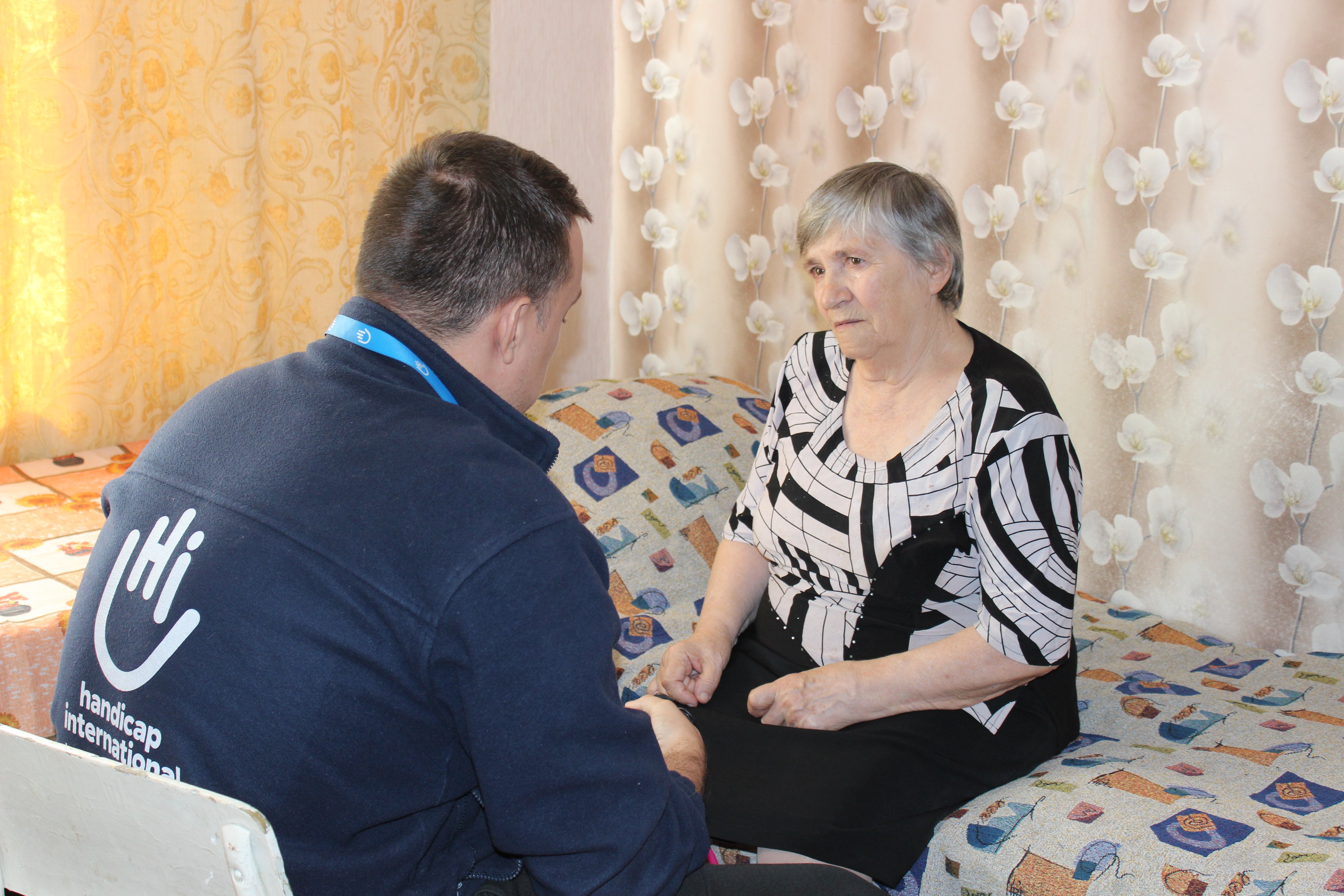 Eine ältere Frau sitzt auf einem Bett und wird von einem HI-Mitarbeiter behandelt. Sie schaut ihn an, ihn sieht man von hinten. 