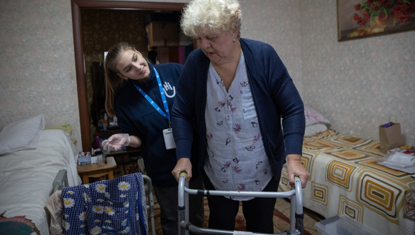 Eine ältere Frau stützt sich auf einen Rollator. Eine junge HI-Mitarbeiterin unterstützt sie. 