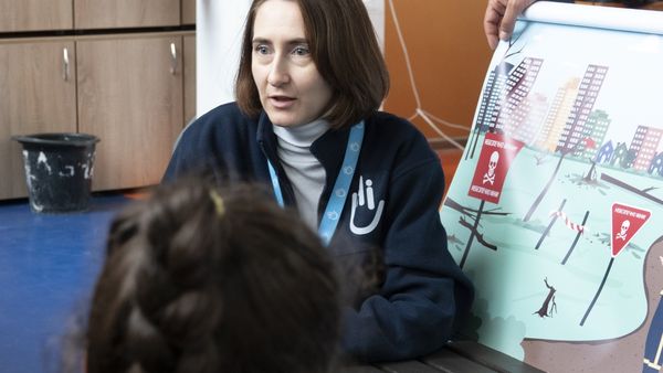 Eine Mitarbeiterin von Handicap International redet und neben ihr steht ein Warnplakat vor Minen und Blindgängern. Im Bild ist noch der Hinterkopf eines Mädchens.