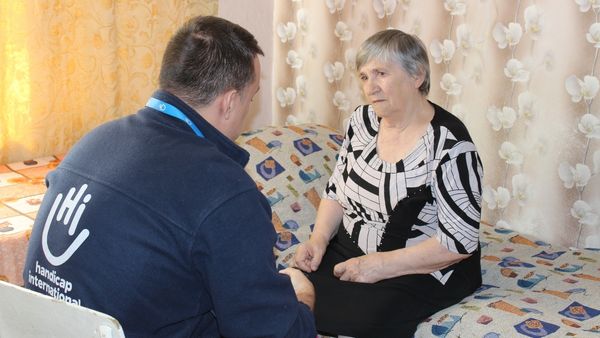 Eine ältere Frau sitzt auf einem Bett und schaut den Mann gegenüber intensiv an. Er trägt eine Weste mit dem Logo von Handicap International.