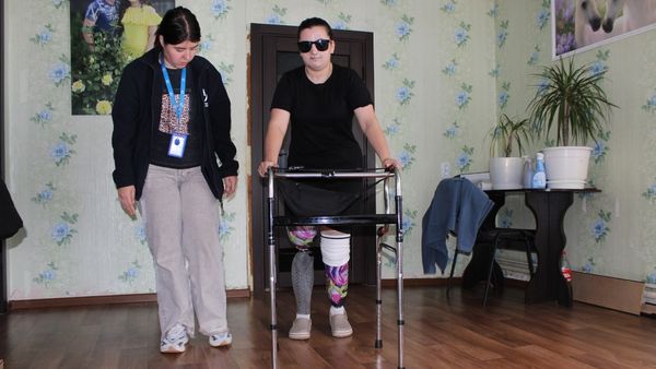 Eine Frau mit Sonnenbrille und zwei Beinprothesen stützt sich auf eine Gehhilfe. Neben ihr begleitet sie eine Mitarbeiterin von Handicap International.