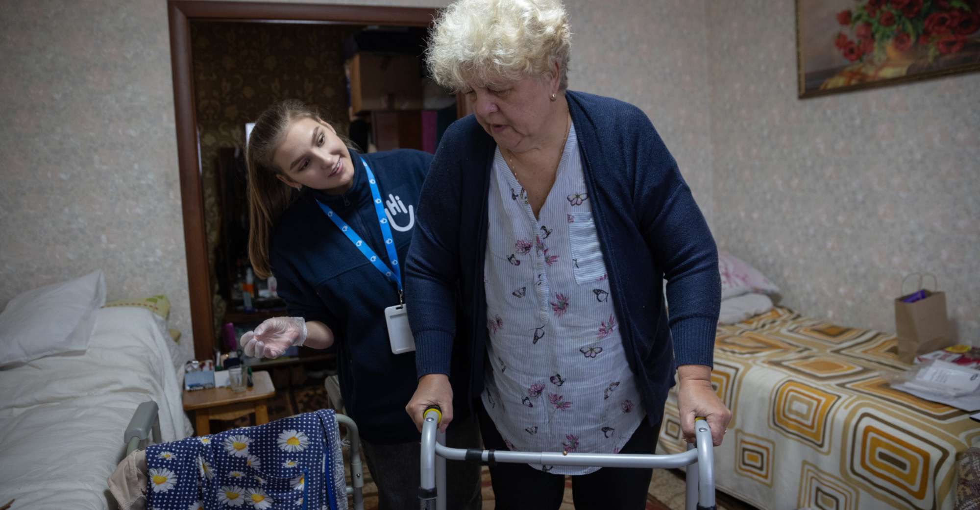 Eine ältere Frau mit kurzen blonden Haaren stützt sich auf ihren Rollator. Hinter ihr steht eine Helferin von Handicap International und lächelt sie an. Im Hintergrund ist ein Wohnzimmer.