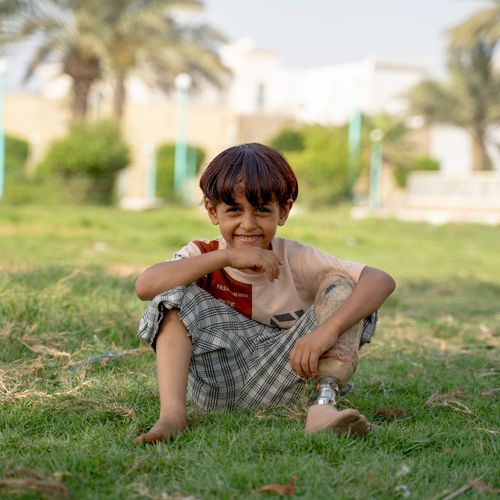 Junge aus dem Jemen mit Beinprothese sitzt auf einer Wiese und lächelt in die Kamera