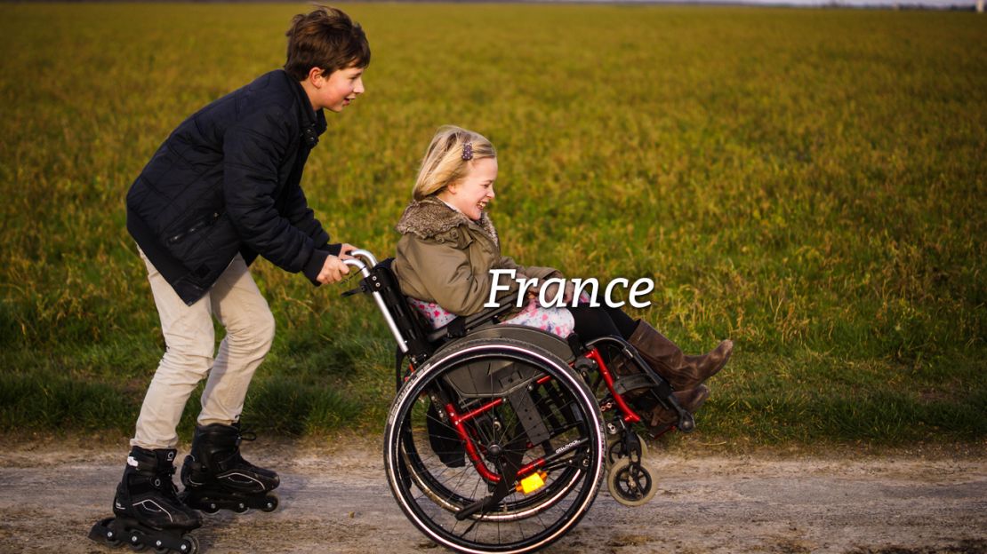 Handicap International Frankreich. Ein gehbehindertes Mädchen im Rollstuhl wird von einem Jungen mit Rollschuhen angeschoben.