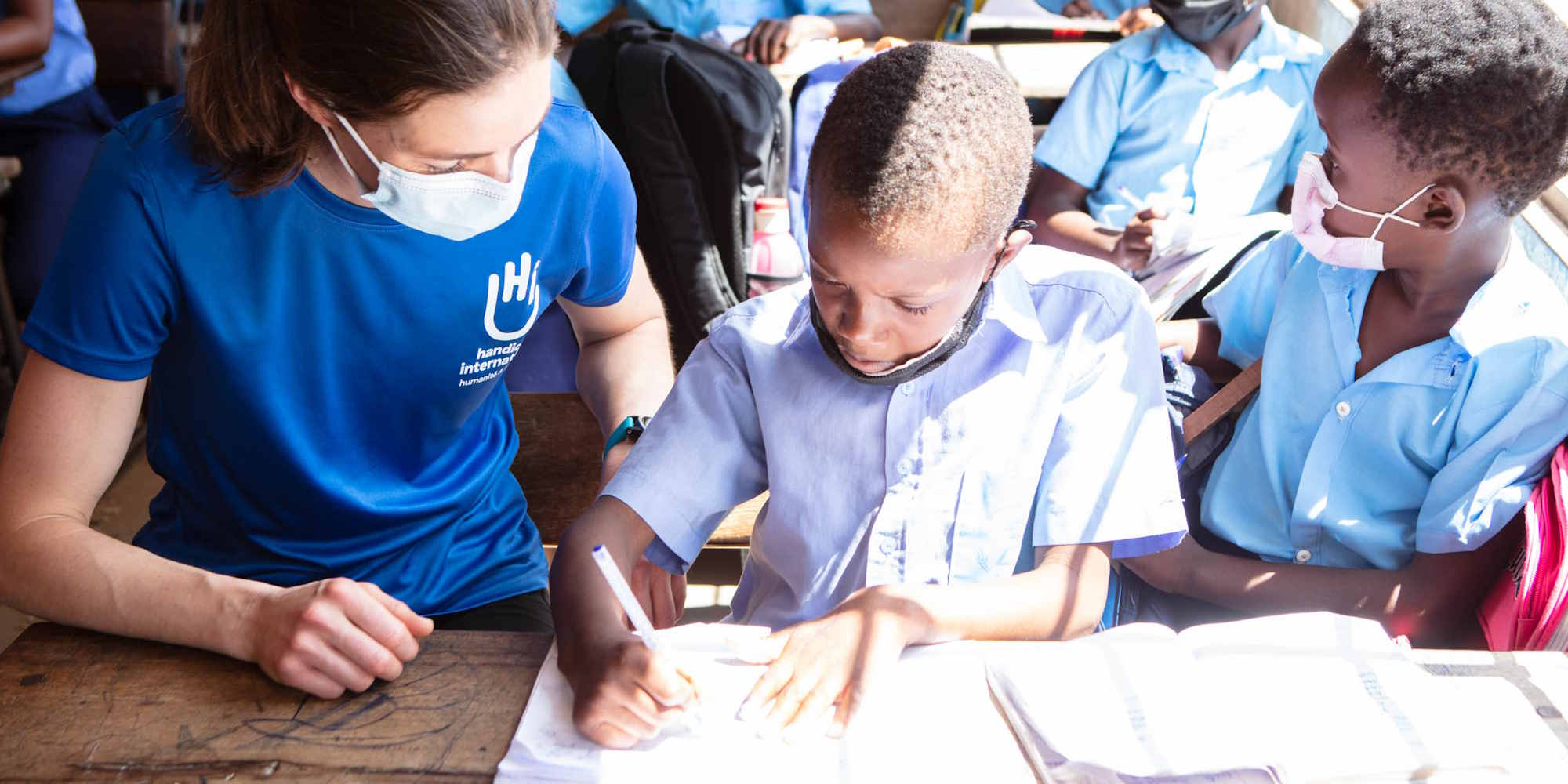 Eine HI-Mitarbeiterin und ein afrikanischer Junge sitzen nebeneinander an der Schulbank, er schreibt konzentriert in sein Schulheft.