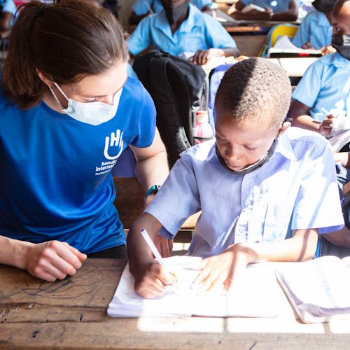 Eine HI-Mitarbeiterin und ein afrikanischer Junge sitzen nebeneinander an der Schulbank, er schreibt konzentriert in sein Schulheft.