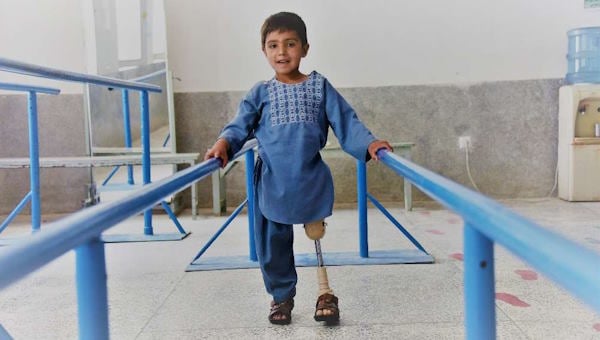 Ein kleiner Junge in afghanischer Kleidung mit einem prosthetischen Bein hält sich an Übungsgeländern fest. Er lächelt.