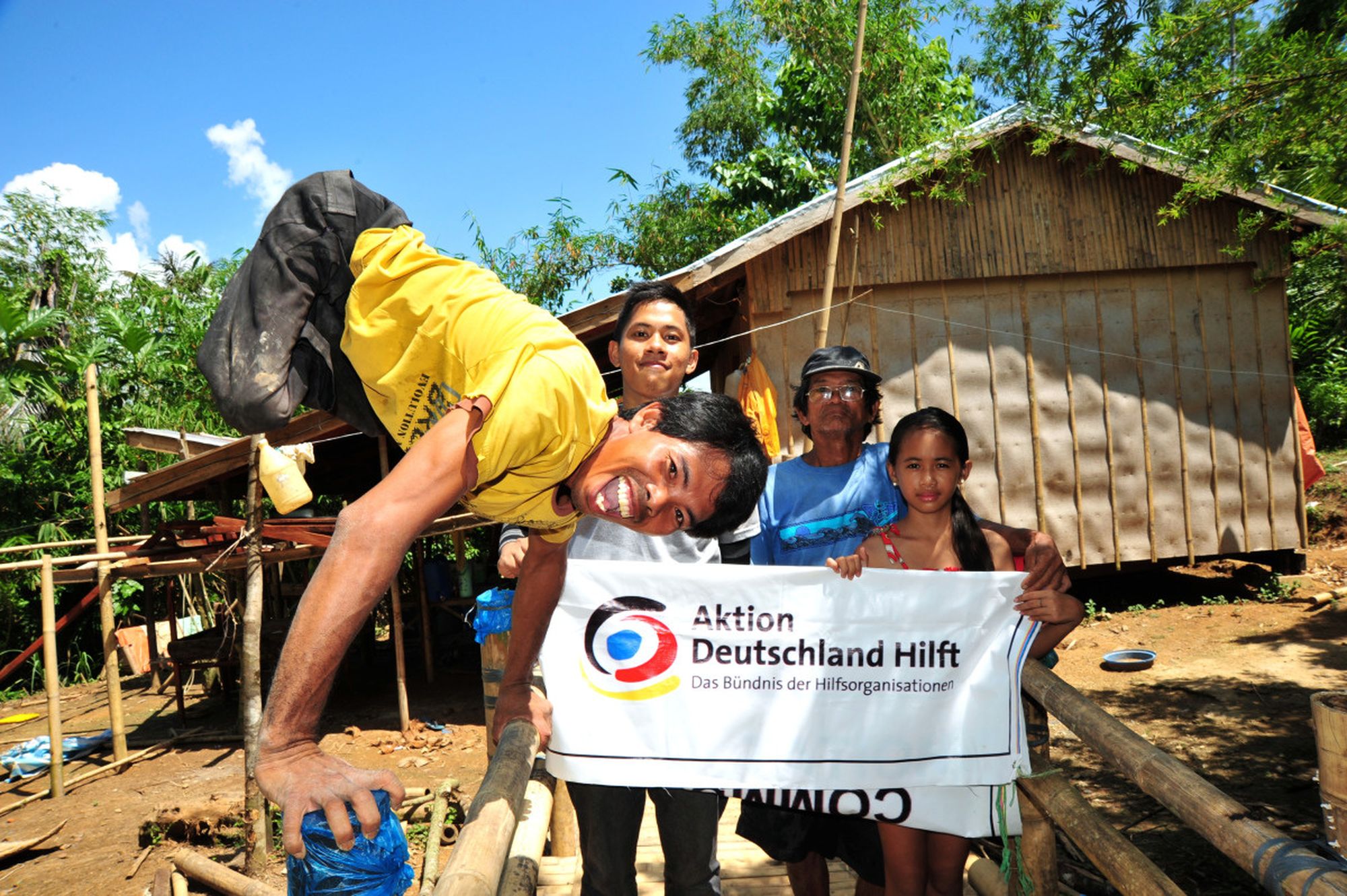 Mann ohne Beine macht einen Handstand und lacht in die Kamera. Im Hintergrund halten Kinder ein Logo-Banner von Aktion Deutschland Hilft hoch