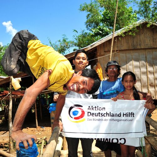 Mann ohne Beine macht einen Handstand und lacht in die Kamera. Im Hintergrund halten Kinder ein Logo-Banner von Aktion Deutschland Hilft hoch