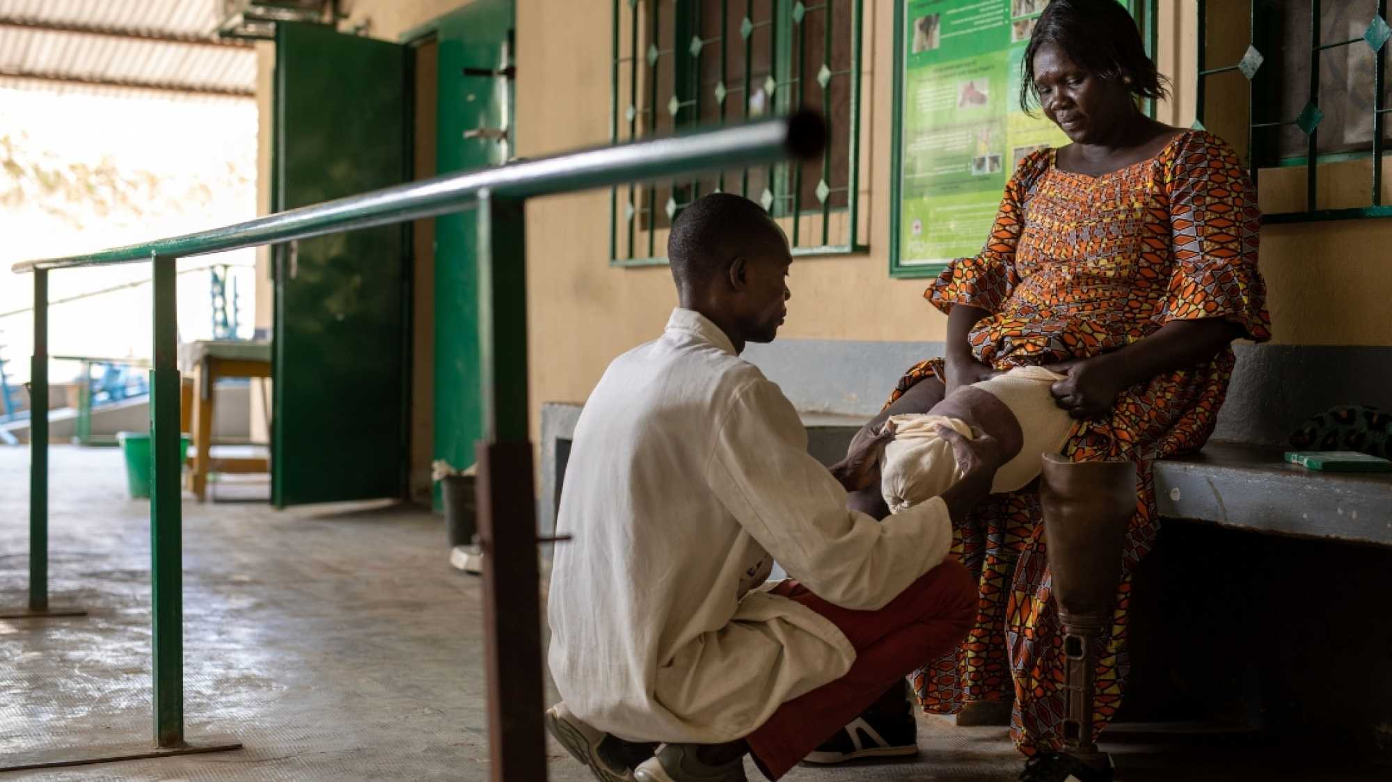 Eine afrikanische Frau sitzt auf einer Bank, ihr links Bein ist ab dem Knie amputiert. Neben ihr steht ihre Beinprothese. Vor ihr kniet ein HI-Mitarbeiter, der ihr Bein untersucht.