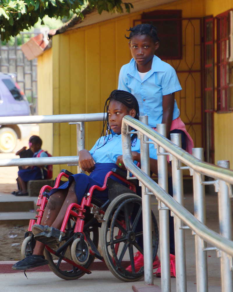 Chelsia, 14 Jahre, sitzt im Rollstuhl und wird von ihrer Freundin eine Rampe zur Schule hochgeschoben